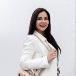 Attractive young woman with a beautiful smile in a fashionable white leather jacket with a gold backpack is standing near the white building on a bright spring day. Positive cute brunette girl model.