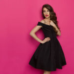 Beautiful young woman in elegant black cocktail dress is holding hand on chest and looking away. Three quarter length studio shot on pink background.