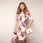 Blonde young woman in floral spring summer dress. Girl posing on a white background. Summer floral outfit. Stylish wavy hairstyle. Fashion photo. Glamour lady with sunglasses and handbag