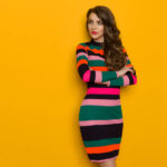 Beautiful young woman in colorful vibrant striped dress with long sleeves is standing with arms crossed and looking away. Three quarter length studio shot on yellow background.