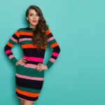 Beautiful young woman in colorful vibrant striped dress with long sleeves is holding hand on hip and looking at camera. Three quarter length studio shot on turquoise background.