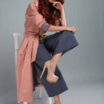 Fashion Model Style. Fashionable Woman In Stylish Clothes Posing On Grey Background In Studio. Beautiful Sexy Girl Wearing Pink Coat, Purple Trousers, Beige Shoes Sitting On Chair. High Resolution